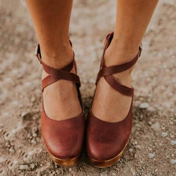 Bruine leren damesschoenen met kruisbanden, comfortabele hak, op een zanderige ondergrond. Perfect voor casual en stijlvolle outfits.