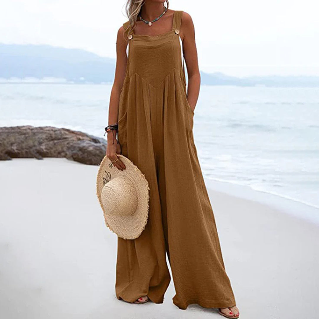 Vrouw in bruine, wijde jumpsuit op strand, houdt strohoed vast. Zomermode, casual strandkleding, comfortabele damesoutfit.