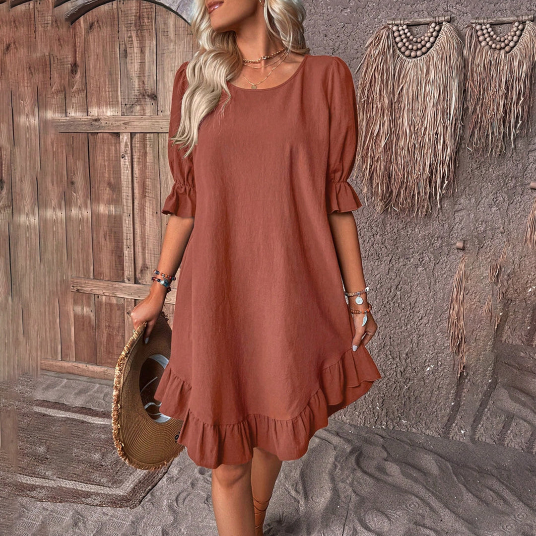 Vrouw in bruine zomerjurk met ruches, staand voor rustieke houten deur. Boho-stijl, casual mode, strandaccessoires, trendy dameskleding.