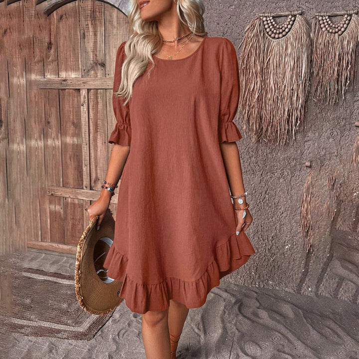 Vrouw in bruine zomerjurk met ruches, staand voor rustieke houten deur. Boho-stijl, casual mode, strandaccessoires, trendy dameskleding.