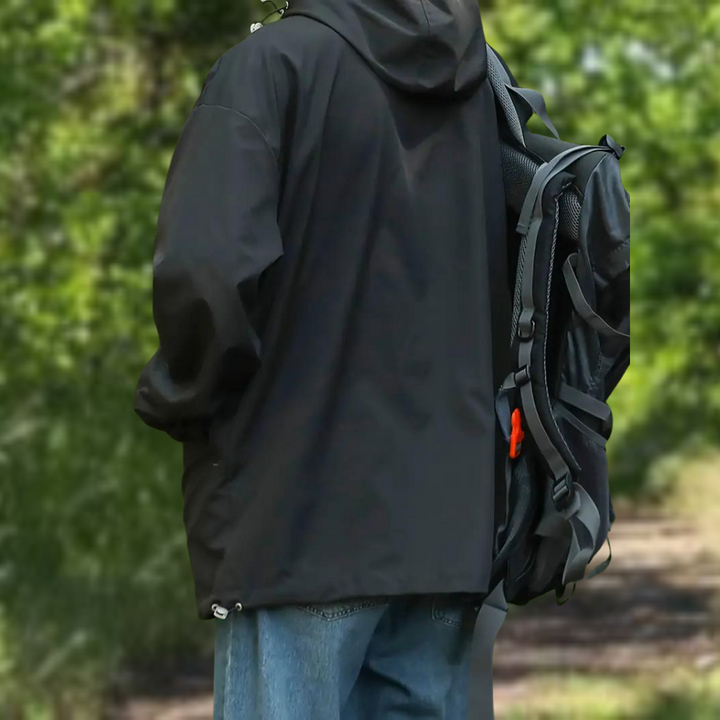 Zwarte waterdichte jas met capuchon, gedragen door persoon met rugzak, buiten in groene omgeving. Ideaal voor wandelen en outdooractiviteiten.