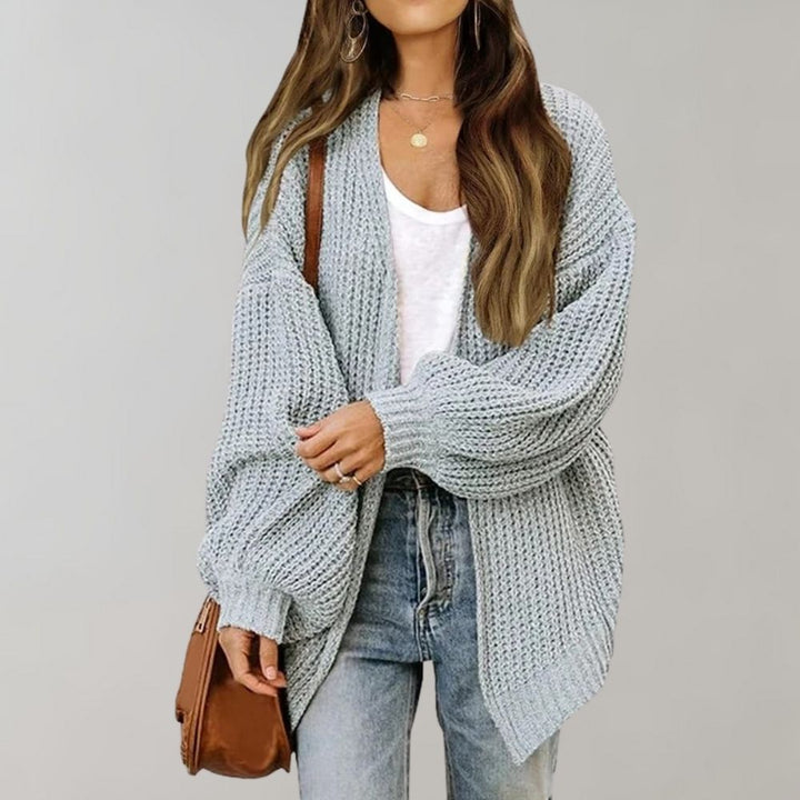 Vrouw in grijze gebreide oversized cardigan, witte top en blauwe jeans, met bruine schoudertas. Modieuze herfstoutfit, casual dameskleding.