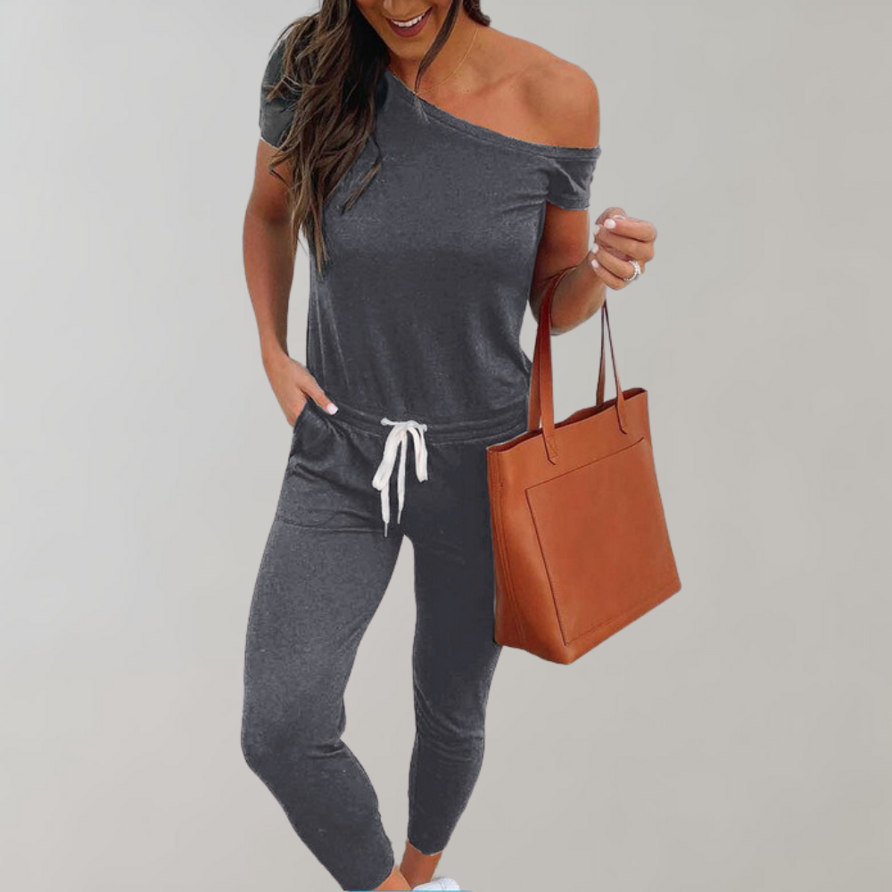 Grijze off-shoulder jumpsuit voor dames met witte trekkoord, gecombineerd met bruine leren handtas. Comfortabele en stijlvolle damesmode.