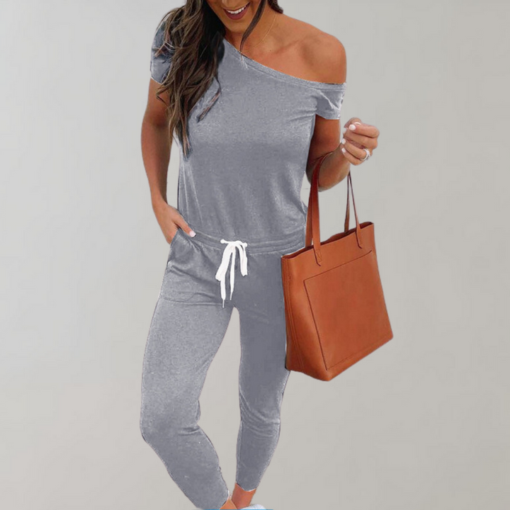 Grijze off-shoulder jumpsuit voor dames, casual stijl, met witte trekkoord en bruine leren handtas. Comfortabele en modieuze outfit.