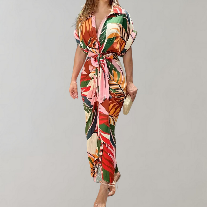 Vrouw in kleurrijke jumpsuit met tropisch bladpatroon, roze ceintuur en clutch. Perfect voor zomermode en stranduitjes.