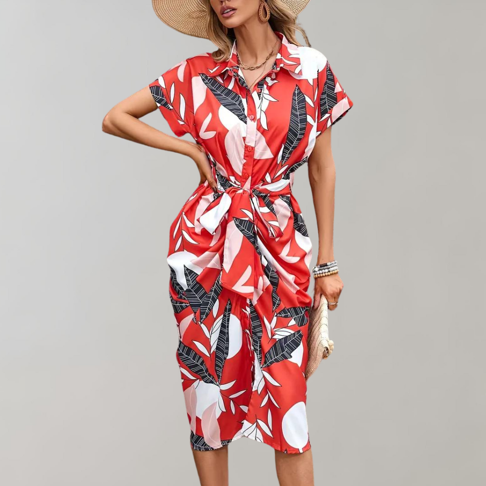 Rode zomerjurk met tropisch bladpatroon, korte mouwen en strikdetail, gedragen door model met hoed. Perfect voor strandmode en casual uitjes.