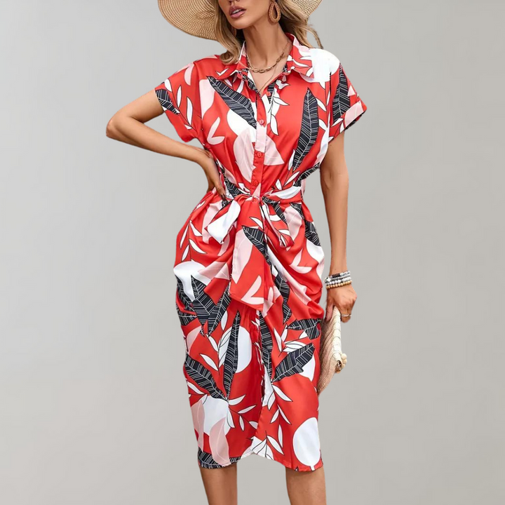 Rode zomerjurk met tropisch bladpatroon, korte mouwen en strikdetail, gedragen door model met hoed. Perfect voor strandmode en casual uitjes.