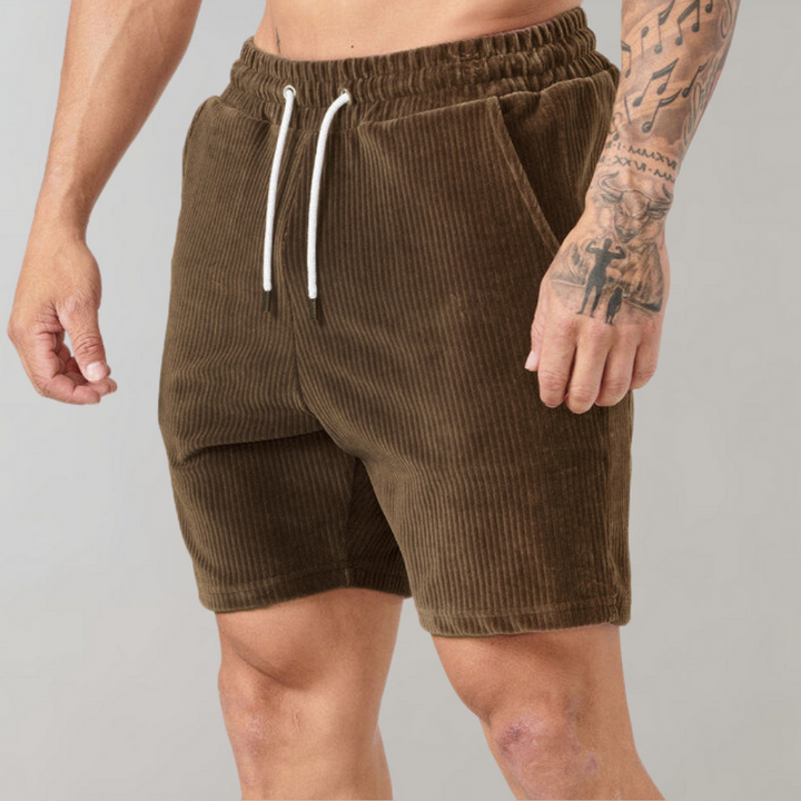 Bruine corduroy herenshorts met elastische tailleband en witte trekkoorden, gedragen door een man met getatoeëerde arm. Comfortabele zomermode.