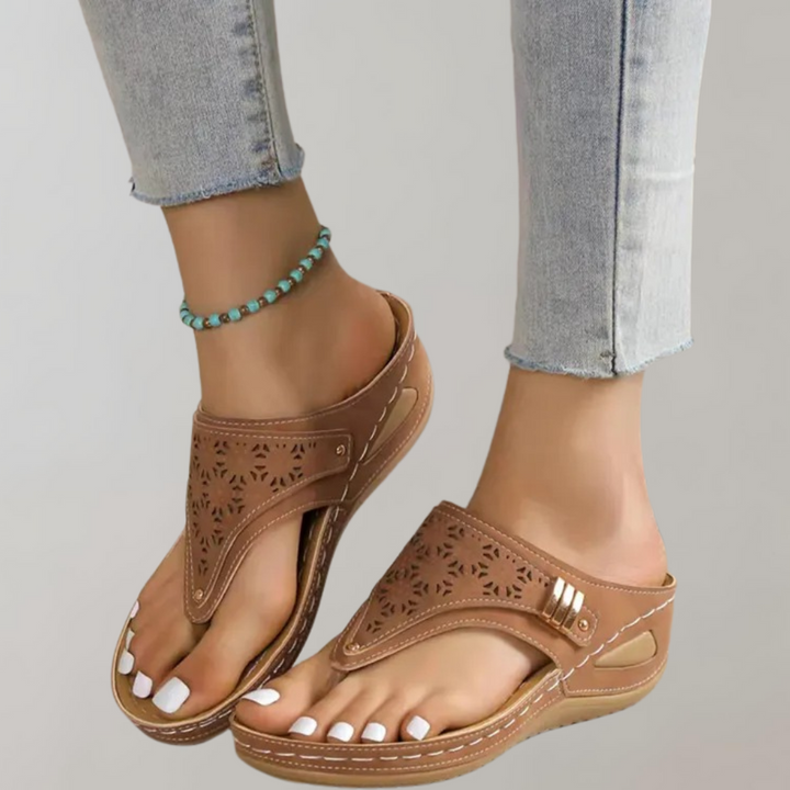 Bruine leren sandalen met uitgesneden patroon, gedragen door vrouw met turquoise enkelbandje en lichtblauwe jeans. Comfortabele zomerschoenen.