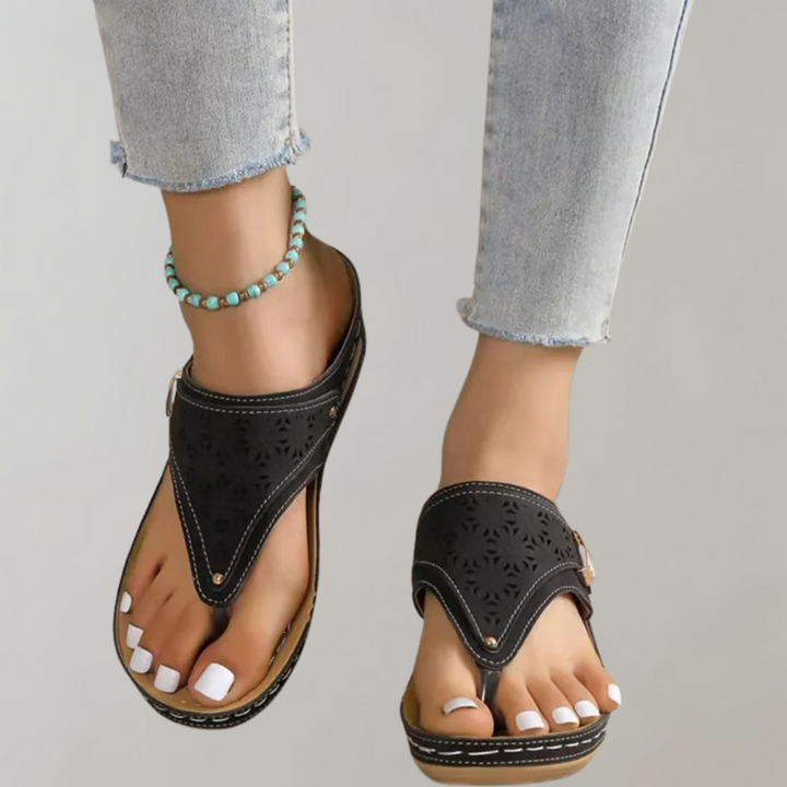 Zwarte leren sandalen met uitgesneden patroon, gedragen door een persoon met witte nagellak en turquoise enkelbandje, op een grijze achtergrond.