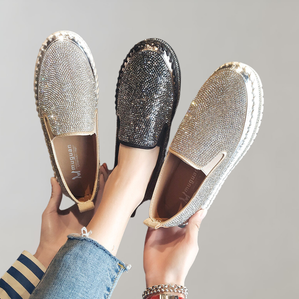 Twee paar glinsterende loafers, zilver en zwart, met strass steentjes, vastgehouden door handen. Modieuze damesschoenen, casual en elegant.
