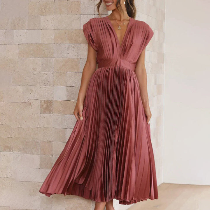 Vrouw in elegante, geplooide roze jurk met V-hals, staand tegen een beige stenen muur. Perfect voor avondkleding of formele evenementen.