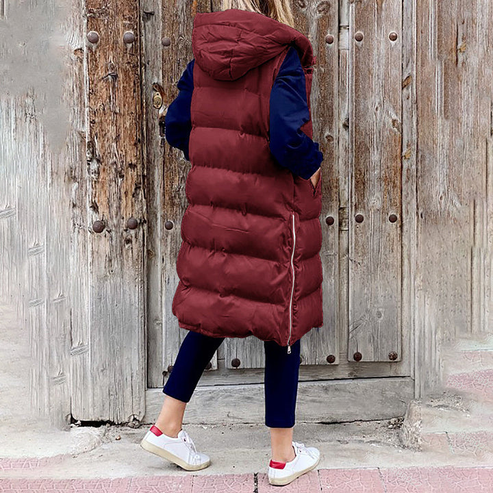 Vrouw in bordeauxrode gewatteerde lange bodywarmer met capuchon, blauwe broek en witte sneakers, staand voor een rustieke houten deur.