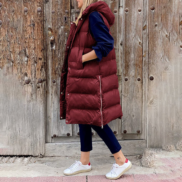 Vrouw in bordeauxrode gewatteerde lange bodywarmer met capuchon, blauwe broek en witte sneakers, voor rustieke houten deur. Wintermode, casual stijl.