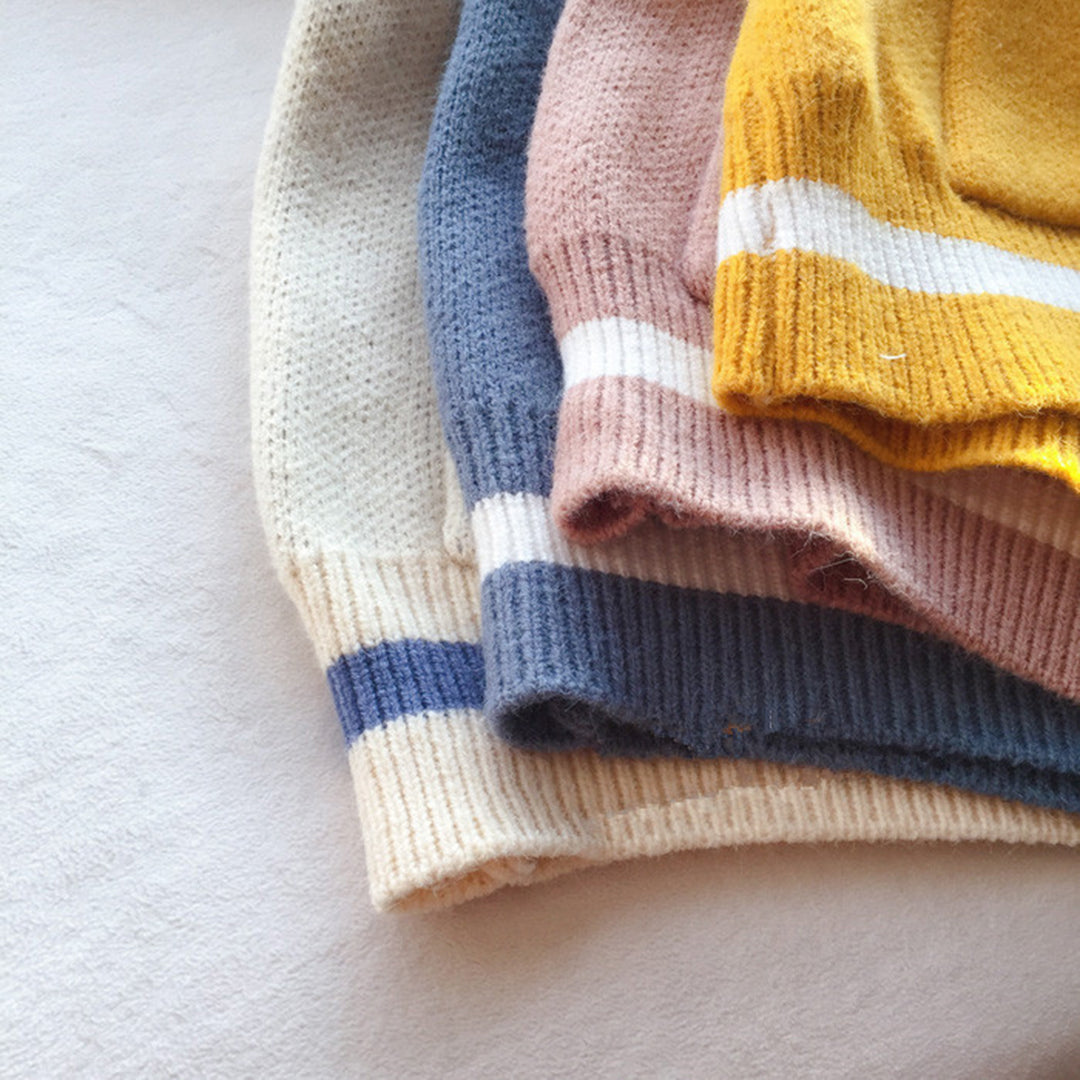 Gestapelde wollen truien in crème, blauw, roze en geel met strepen, liggend op een witte achtergrond. Zachte, warme kleding voor herfst en winter.