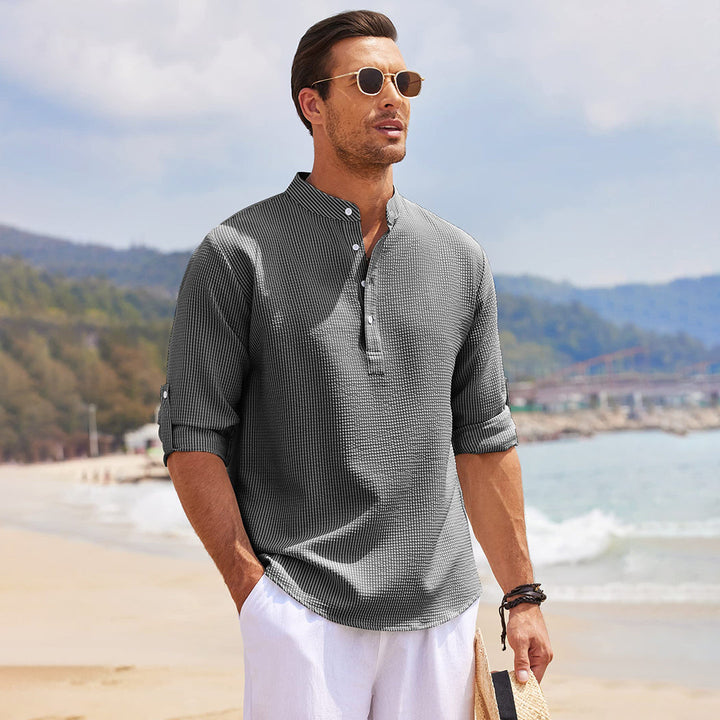 Man in grijs casual overhemd met opgerolde mouwen op strand, draagt zonnebril. Stijlvolle herenkleding, zomermode, strandoutfit.