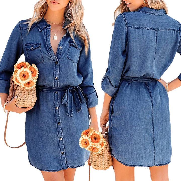Blauwe denim jurk met lange mouwen, knoopsluiting en ceintuur, gedragen door model met rieten tas en oranje bloemen. Casual damesmode.