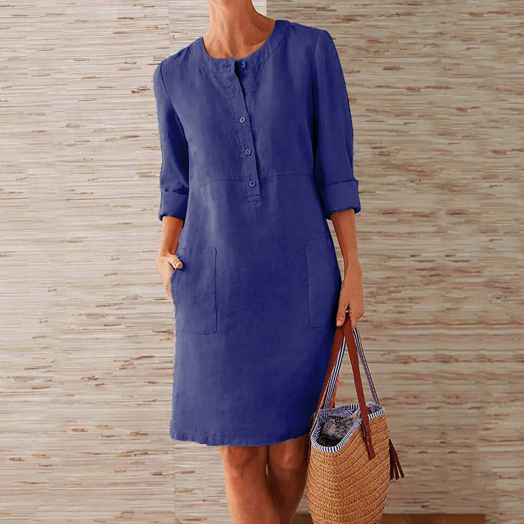 Donkerblauwe linnen jurk met knoopsluiting en opgestroopte mouwen, gedragen door vrouw met rieten tas. Casual damesmode, zomerjurk.