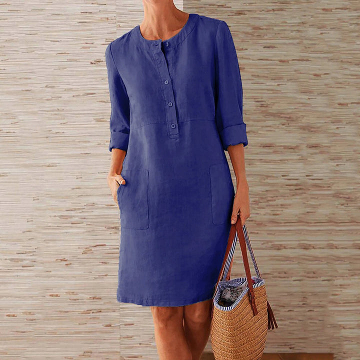 Donkerblauwe linnen jurk met knoopsluiting en opgestroopte mouwen, gedragen door vrouw met rieten tas. Casual damesmode, zomerjurk.