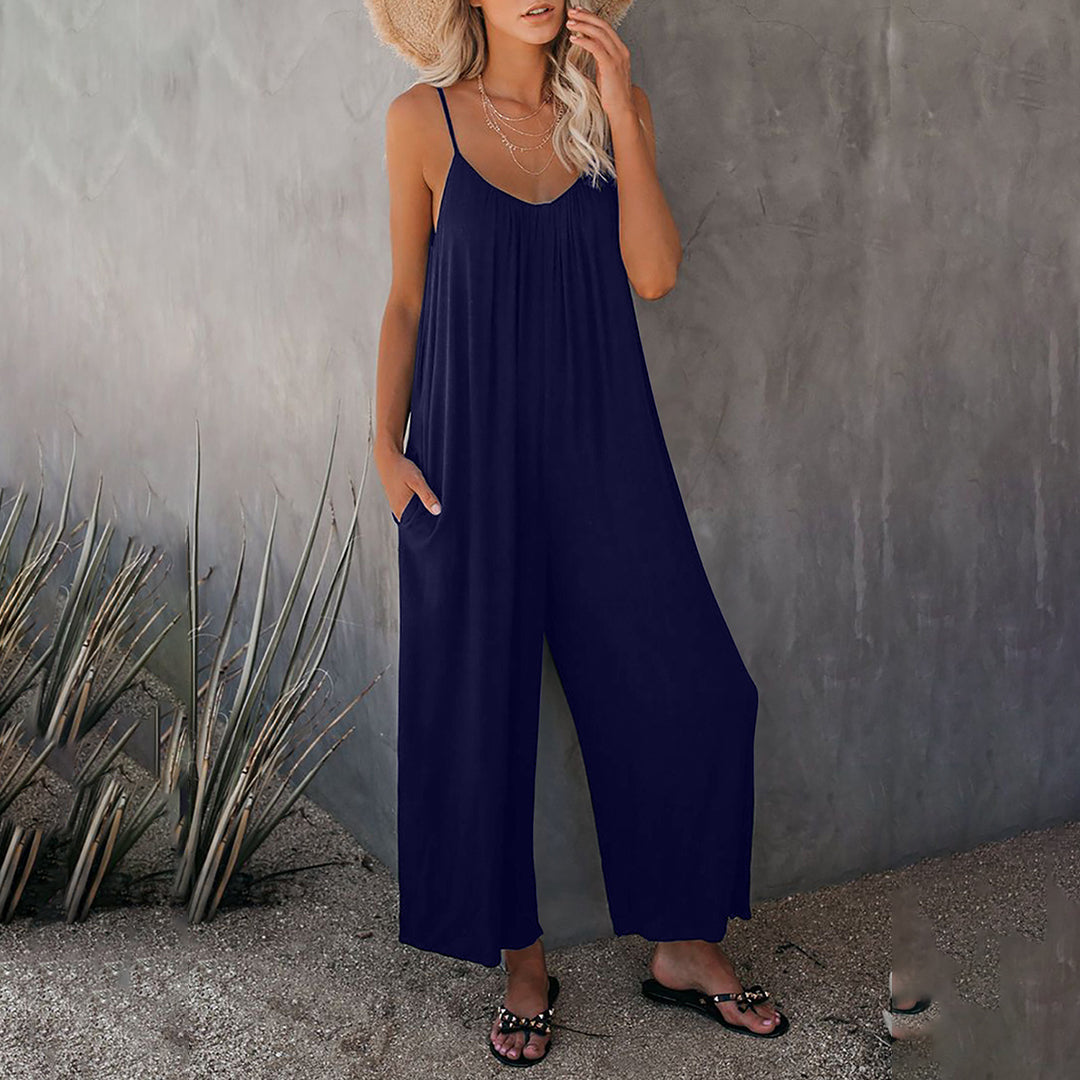 Vrouw in stijlvolle donkerblauwe jumpsuit met spaghetti bandjes, zomerse hoed en sandalen, poseert tegen een betonnen muur. Perfect voor casual zomeroutfits.