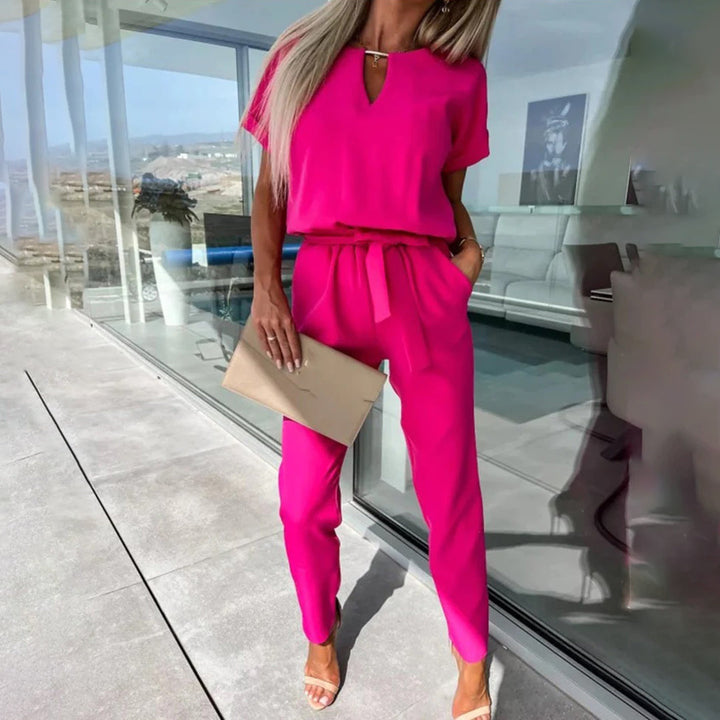 Vrouw in fuchsia jumpsuit met korte mouwen en strikceintuur, staand op terras met glazen wand. Ze houdt een beige clutch vast. Zomermode, elegante stijl.