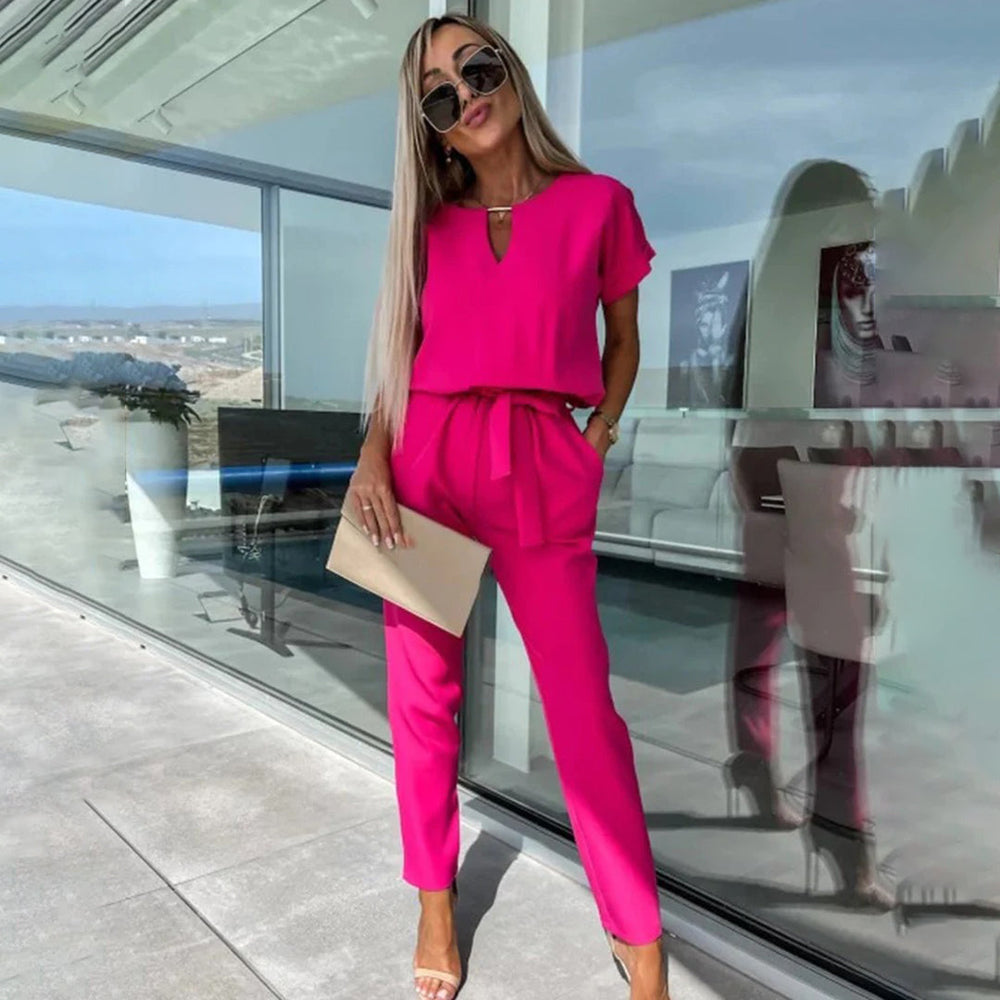 Vrouw in fuchsia jumpsuit met zonnebril en enveloptas, staand voor glazen wand. Trendy damesmode, zomeroutfit, stijlvolle kleding.
