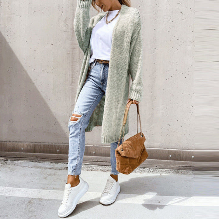 Vrouw in casual outfit met grijze lange cardigan, witte sneakers, gescheurde jeans en bruine tas tegen betonnen achtergrond. Modieuze dameskleding.
