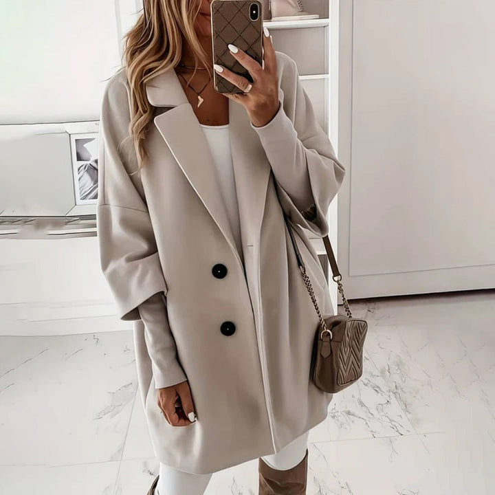 Vrouw in stijlvolle grijze oversized jas met zwarte knopen, witte outfit en beige schoudertas, modieuze herfstkleding, binnenshuis.