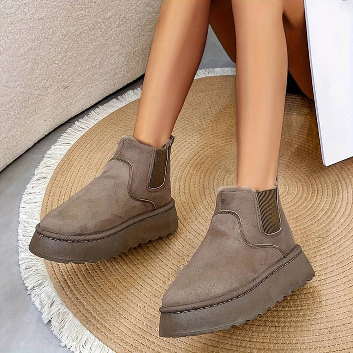Grijze suède enkellaarsjes met dikke plateauzool op een ronde beige mat. Trendy damesmode, comfortabele winterlaarzen, casual stijl.