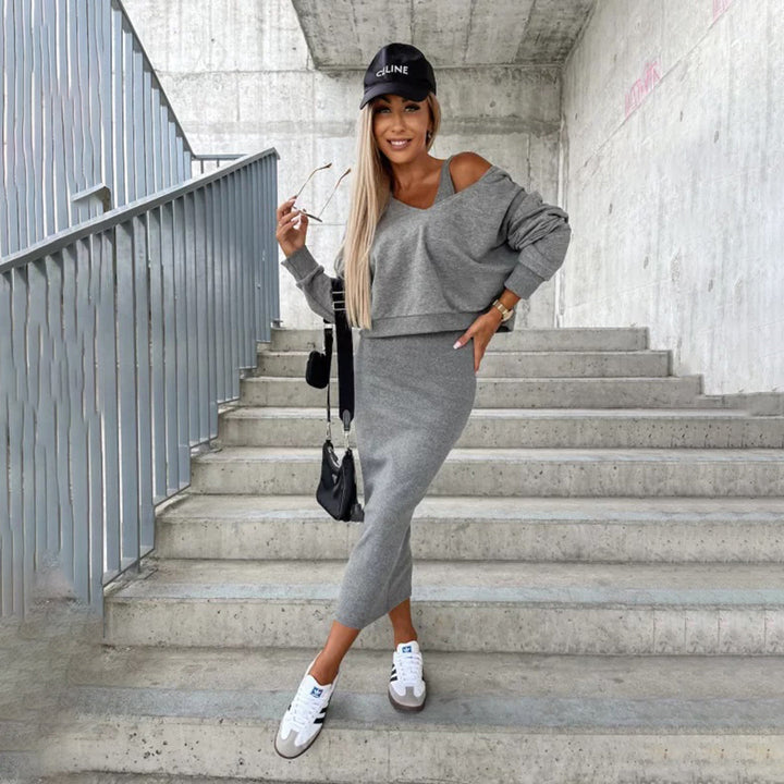 Vrouw in grijze off-shoulder trui en rok, zwarte pet en sneakers, poseert op betonnen trap. Modieuze casual outfit, stedelijke achtergrond.