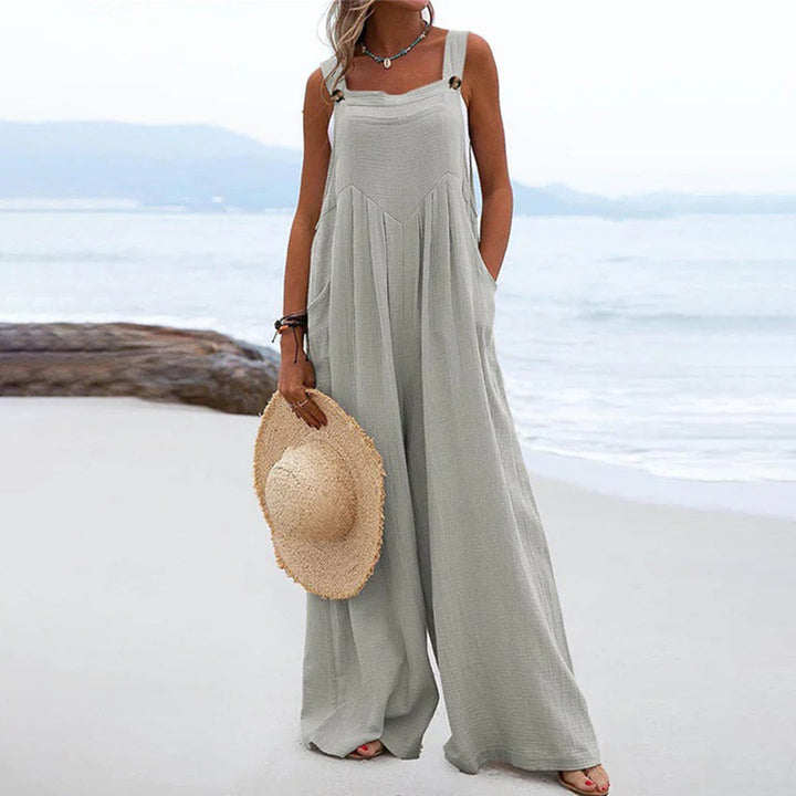 Vrouw in grijze, wijde jumpsuit op strand met strohoed; zomermode, casual strandkleding, comfortabele damesoutfit.