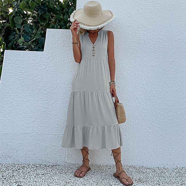 Vrouw in grijze zomerjurk met knopen, strohoed en sandalen, poseert tegen witte muur. Perfecte outfit voor zomerse dagen.