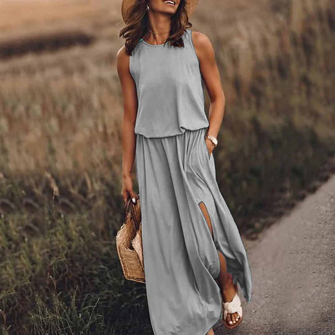Vrouw in grijze maxi-jurk met hoge split, strohoed en rieten tas, wandelend op landweg. Zomerjurk, casual mode, dameskleding.