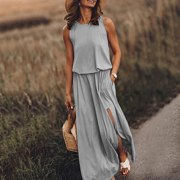 Vrouw in grijze maxi-jurk met hoge split, strohoed en rieten tas, wandelend op landweg. Zomerjurk, casual mode, dameskleding.