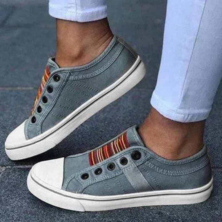 Grijze casual sneakers zonder veters, met kleurrijke strepen en witte zolen, gedragen door persoon in witte broek. Comfortabele en modieuze schoenen.