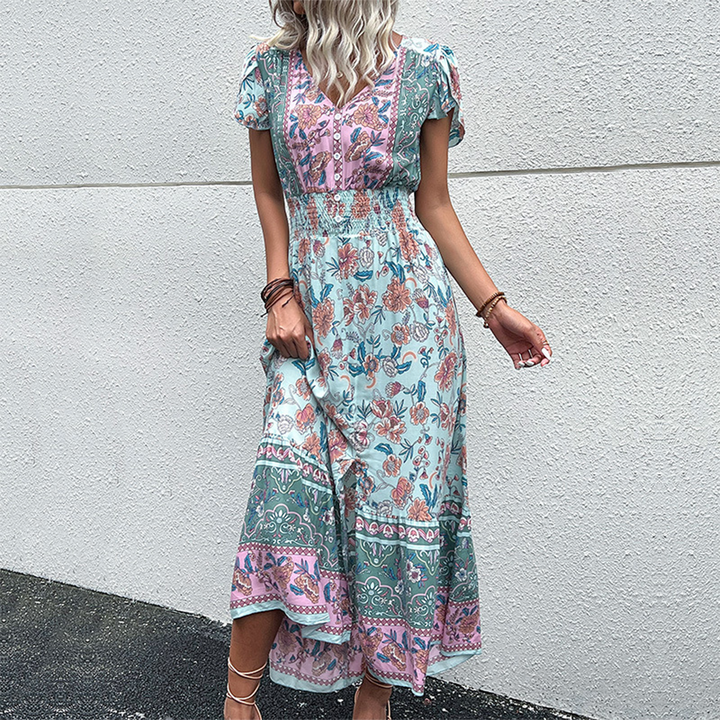 Vrouw in lange, groene bohemian jurk met bloemenprint en korte mouwen, staand tegen een witte muur. Perfect voor zomermode en casual stijl.