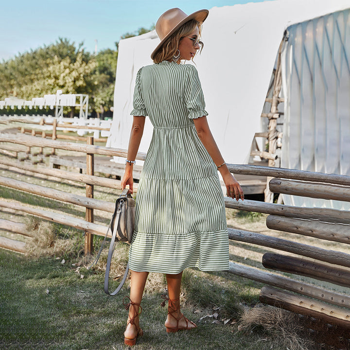 Vrouw in groene gestreepte zomerjurk met hoed en tas, staand bij houten hek in landelijke omgeving. Zomermode, casual dameskleding, buitenstijl.