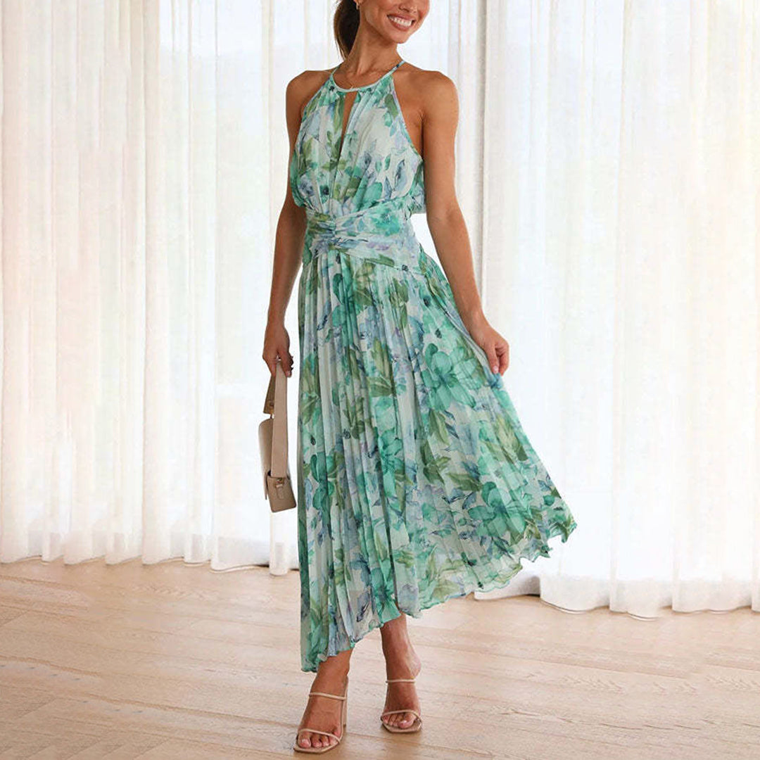 Vrouw in elegante groene zomerjurk met bloemenprint, asymmetrische zoom en halternek, poseert in lichte kamer. Perfect voor zomermode en casual chic.