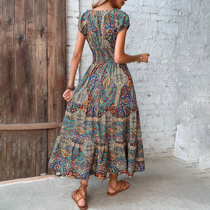Vrouw in kleurrijke bohemian maxi-jurk met paisley-patroon, staand voor houten deur en witte bakstenen muur. Zomerse mode, boho-stijl.