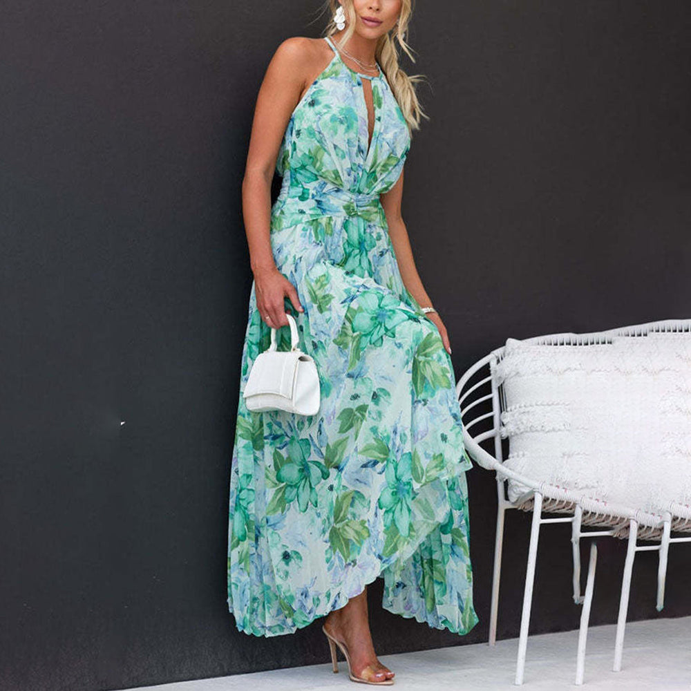 Vrouw in elegante groene bloemenjurk met halternek, witte handtas en hoge hakken, leunend tegen een zwarte muur naast een witte stoel.
