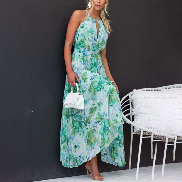 Vrouw in elegante groene bloemenjurk met halternek, witte handtas en hoge hakken, leunend tegen een zwarte muur naast een witte stoel.