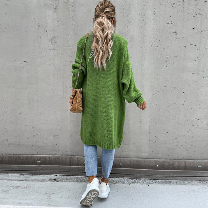 Vrouw in groene oversized trui, blauwe jeans en witte sneakers, met beige schoudertas, tegen een betonnen muur. Modieuze herfstoutfit.