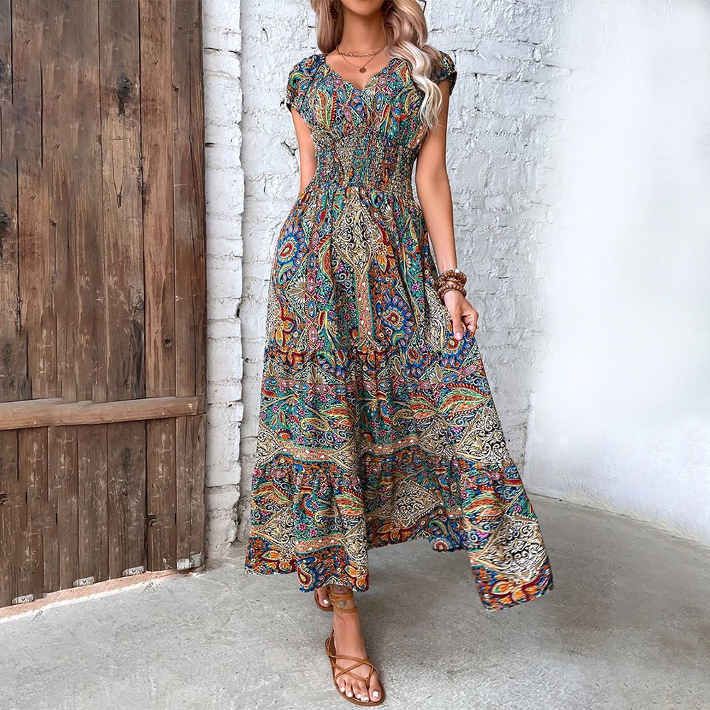 Vrouw in kleurrijke bohemian maxi-jurk met paisley-patroon, staand voor houten deur en witte bakstenen muur. Zomermode, boho-stijl, casual elegantie.