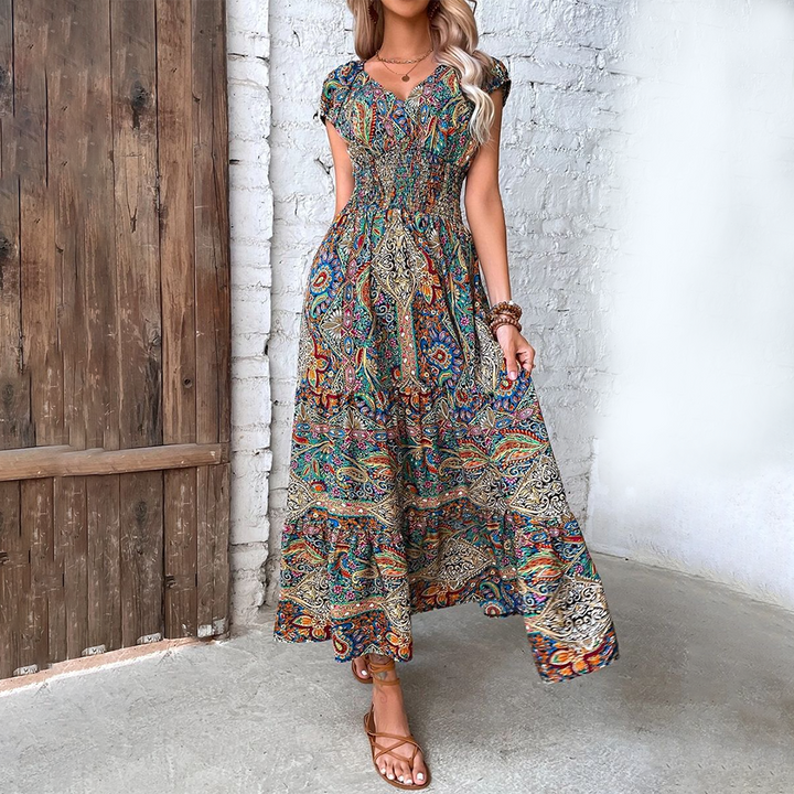 Vrouw in kleurrijke bohemian maxi-jurk met paisley-patroon, staand voor houten deur en witte bakstenen muur. Zomermode, boho-stijl, casual elegantie.