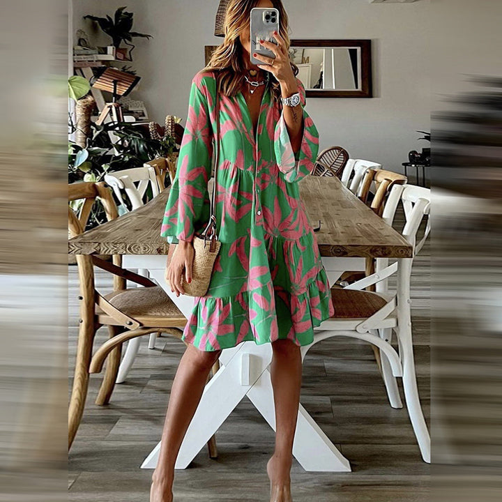 Vrouw in groene en roze bloemenjurk, staand in moderne eetkamer met houten tafel en witte stoelen, modieuze zomeroutfit, casual stijl.