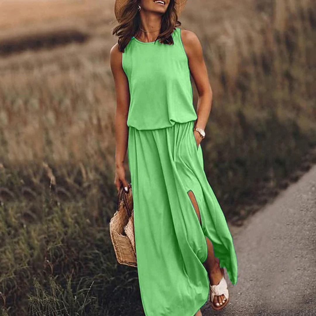 Vrouw in groene maxi-jurk met hoge split, draagt strohoed en rieten tas, wandelend langs een landelijk pad. Zomerjurk, casual mode.