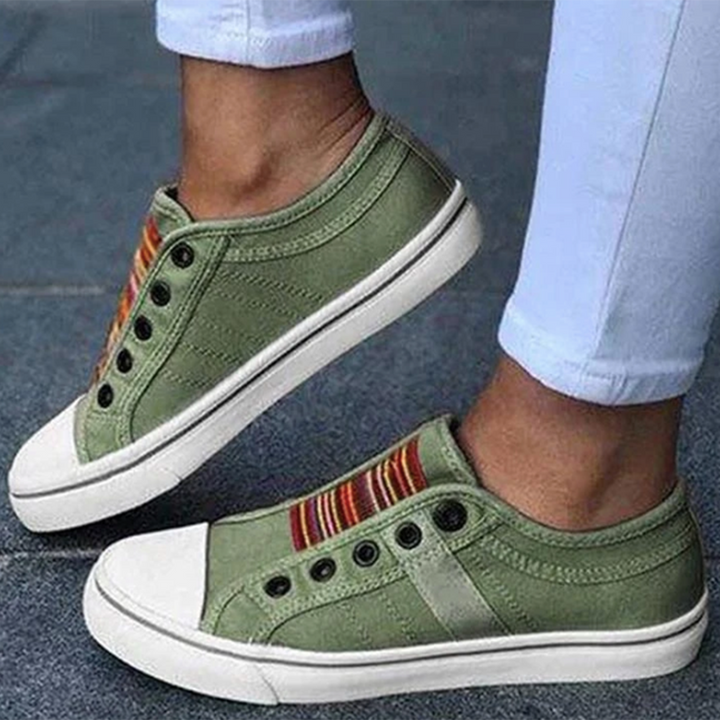 Groene canvas sneakers zonder veters, met kleurrijke strepen en witte zolen, gedragen door persoon in witte broek. Casual en trendy schoenen.