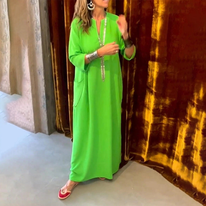 Vrouw in felgroene kaftan met lange mouwen en zakken, staand voor een fluwelen bruine achtergrond. Trendy damesmode, comfortabele kledingstijl.
