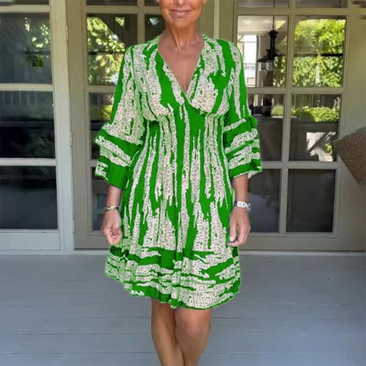 Vrouw in groene zomerjurk met witte print, staand voor glazen deuren. Modieuze dameskleding, casual stijl, comfortabele pasvorm.