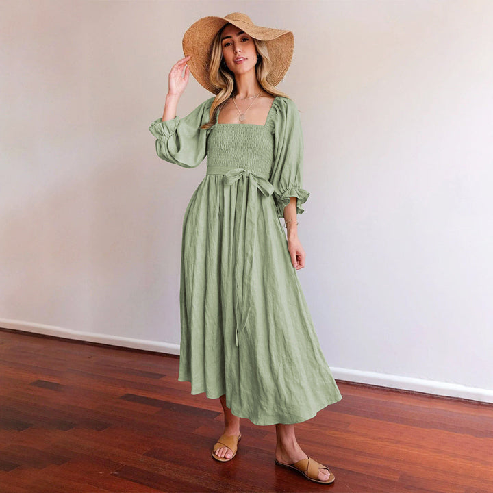 Vrouw in groene, lange jurk met strohoed, staand op houten vloer. Zomerse mode, casual elegantie, dameskleding, boho-stijl.